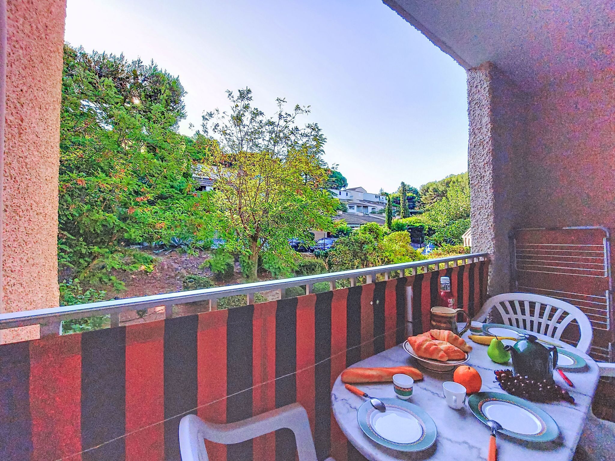 A balcony with a table set for dining, overlooking a garden with trees and a building in the background.