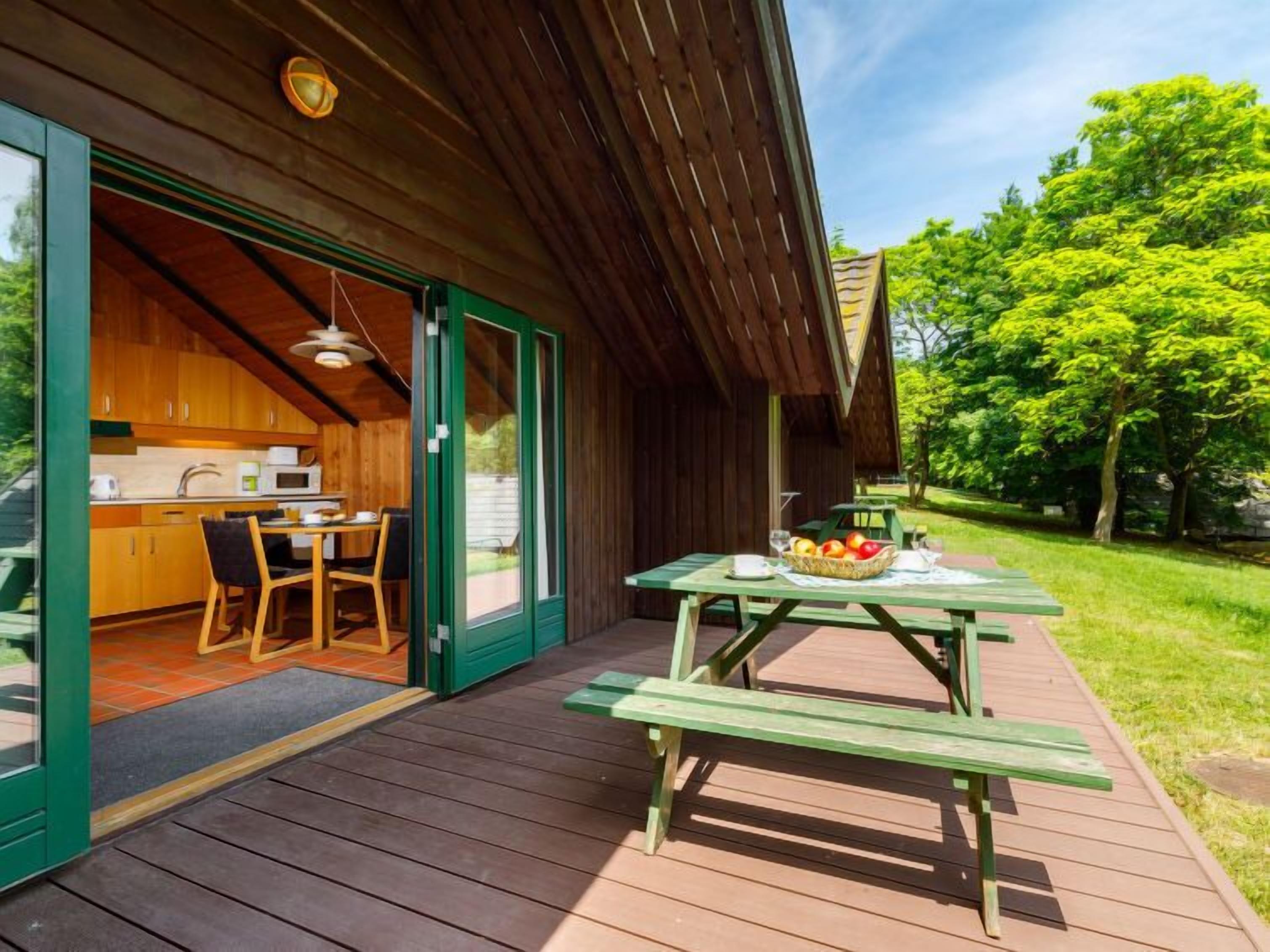 A wooden cabin with a kitchen and dining area visible through glass doors, a picnic table with fruit, and a grassy outdoor area.