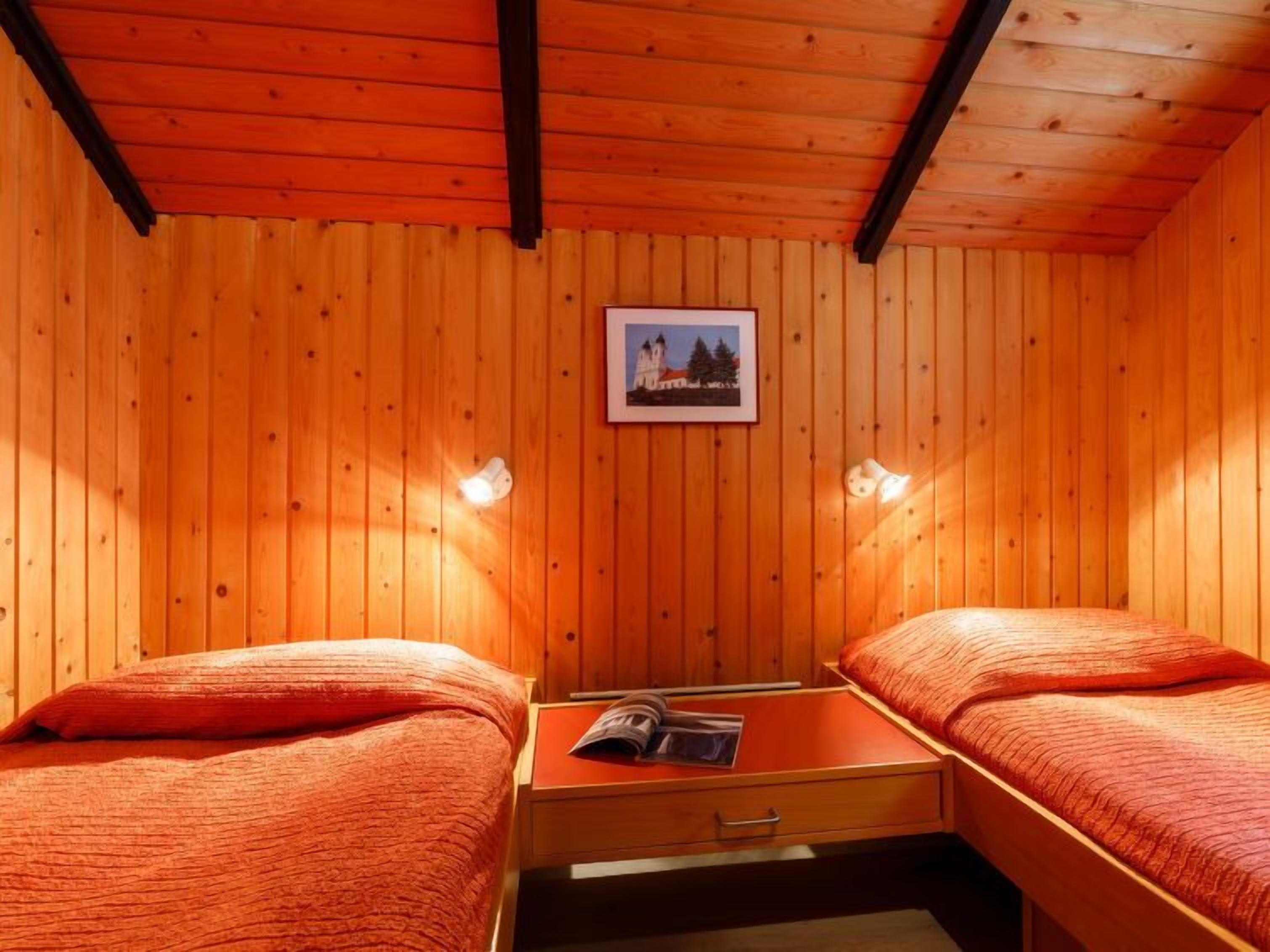 A room with wooden walls and a wooden ceiling. There are two beds with red bedding, a small table with a magazine, and a framed picture on the wall.