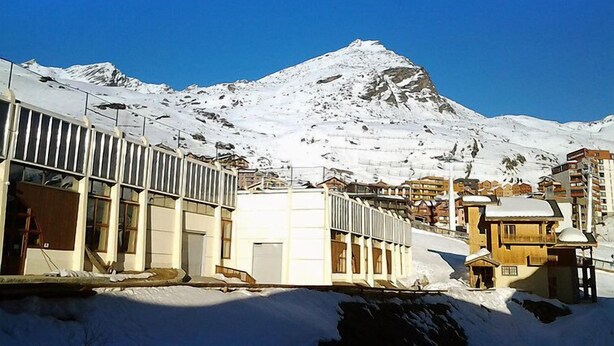 Ein schneegebdeckter Berg mit wolkenlosem, blauem Himmel. Es sind Gebäude mit großen Fenstern und eine Holzhütte mit verschneitem Dach zu sehen.