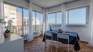 Een eetruimte met een tafel voor vier personen, grote ramen en uitzicht op gebouwen en de zee.