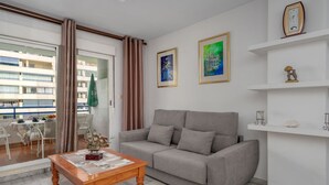 Ein Wohnzimmer mit grauem Sofa, einem Holztisch und einem Balkon mit Blick auf ein Gebäude.