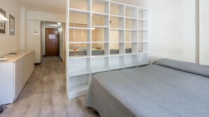 Ein modernes Schlafzimmer mit einem grauen Bett, einem weißen Kleiderschrank mit vielen Regalen, einem Schreibtisch mit Stuhl und einem durch eine Glastür einsehbaren Badezimmer.