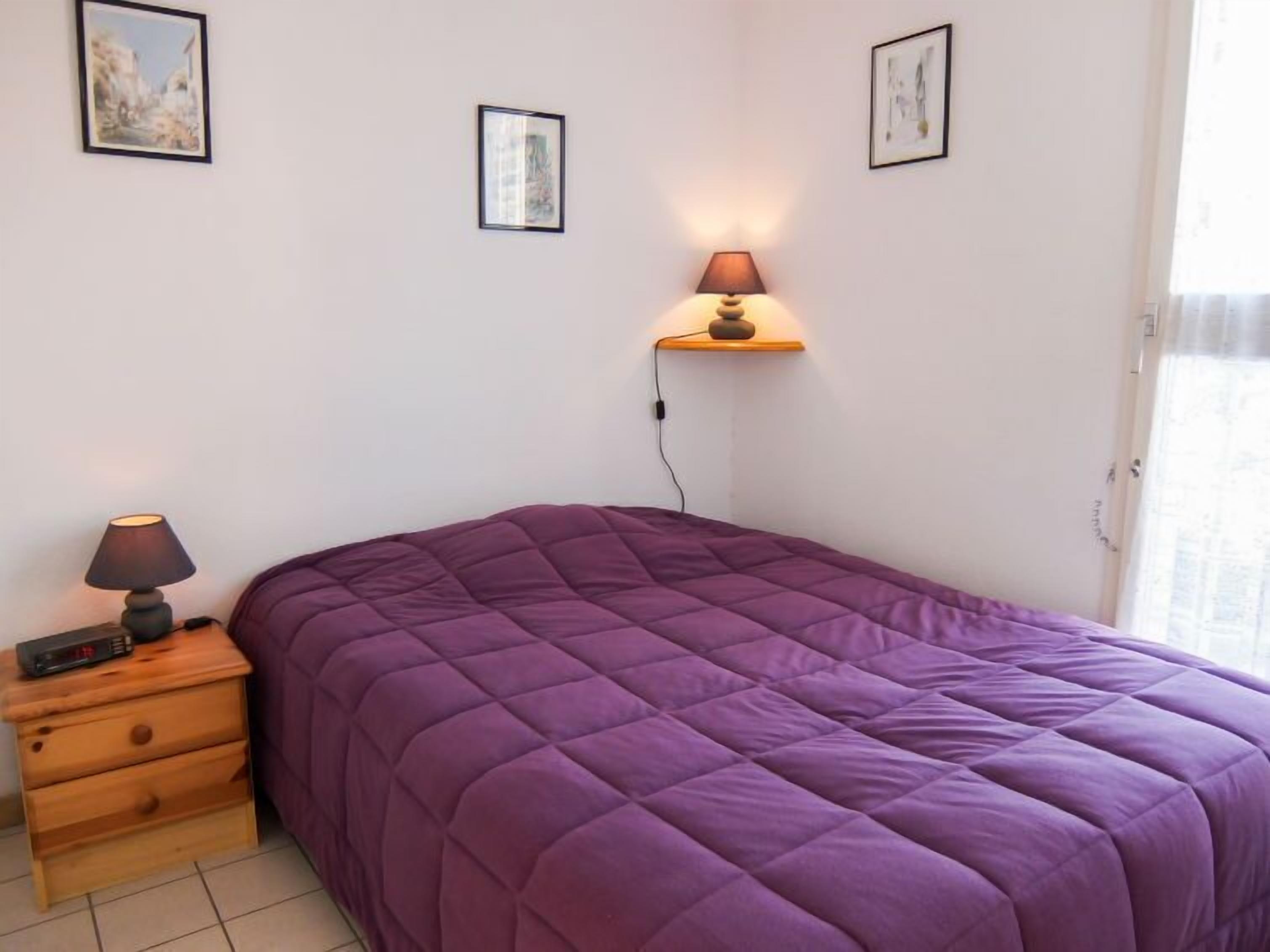 A bedroom with a purple bedspread, a bedside table with a lamp, and two framed pictures on the wall.