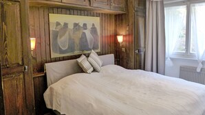 A bedroom with a wooden headboard, a bed with white linens, two wall-mounted lamps, and a window with curtains.