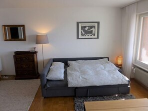 Un soggiorno moderno con un divano grigio, due cuscini bianchi, un settimino in legno, una lampada da terra e un quadro incorniciato appeso al muro.
