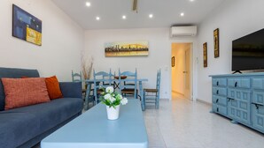 A living room with a blue sofa, a coffee table with flowers, a dining area with blue chairs, and a TV on the right. - Gladiols by Interhome (Cambrils)