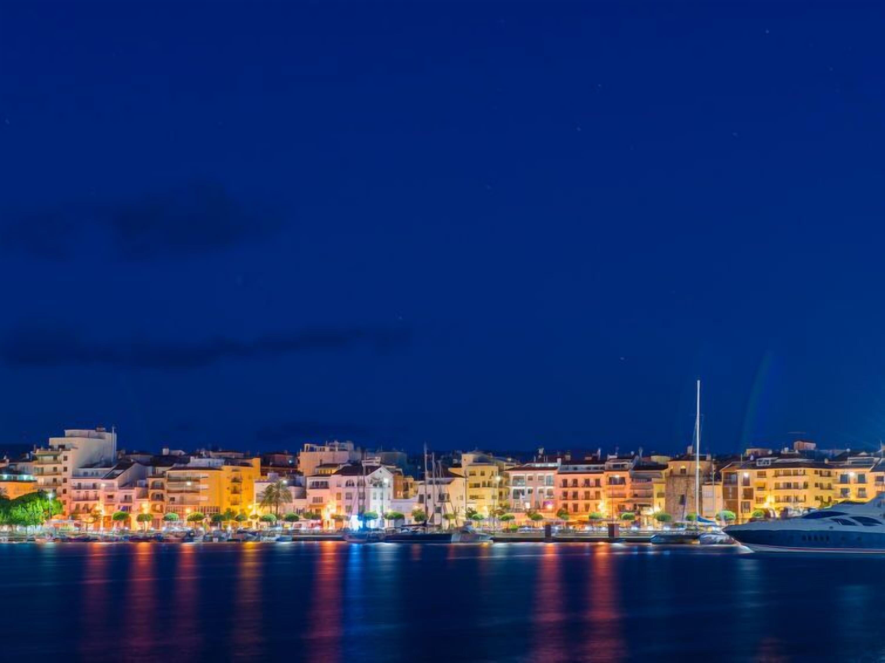 Une vue nocturne sur un paysage urbain le long du littoral, avec des bâtiments illuminés et un yacht amarré au premier plan.