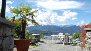An outdoor patio with a stone floor, a large potted palm tree, white chairs, and a table. In the background, there is a view of mountains and a lake. - Vacation home Ursula by Interhome (Pugerna)