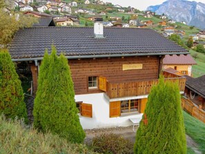 A wooden chalet with a sloped roof, surrounded by coniferous trees, set in a mountainous village.