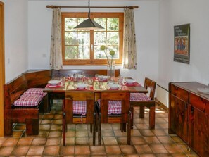 A dining area with a wooden table set for four, surrounded by wooden chairs with checkered cushions. A window with curtains allows natural light to enter, and a poster is visible on the wall.
