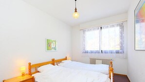 A simple, clean bedroom with a wooden bed, bedside lamps, a radiator, and a window with sheer curtains. - Marignac 33 by Interhome (Villars)