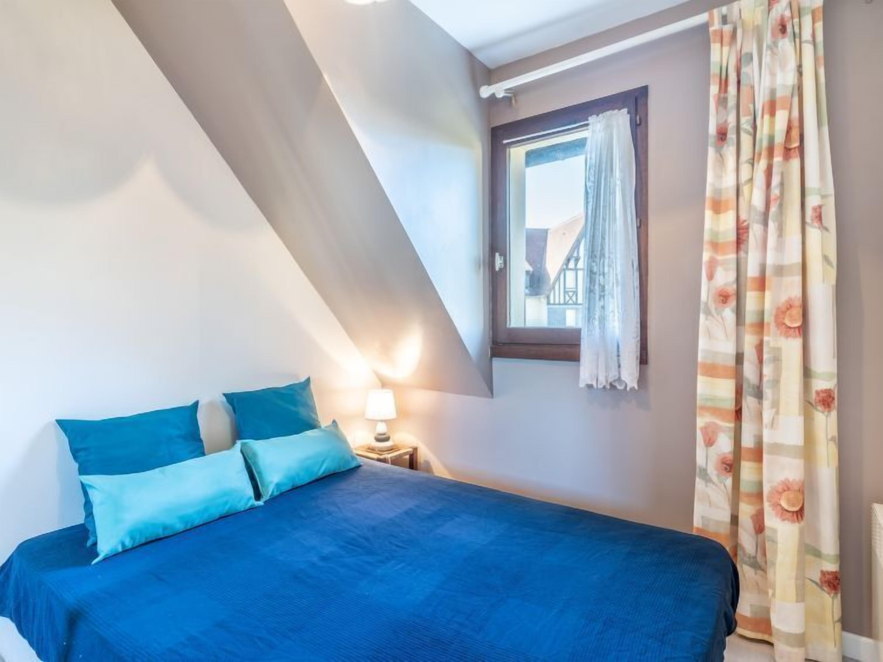 Een slaapkamer met een bed, een nachtkastje, een lamp, een raam met gordijnen en uitzicht op gebouwen.