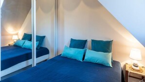 A bedroom with two beds, each with blue bedding and teal pillows. There is a nightstand with a lamp and a decorative item on it. - Fleur Marine by Interhome (Cabourg)