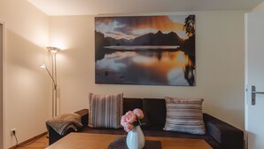 A modern living room with a wooden dining table, a sofa with striped cushions, a vase with flowers, and a large landscape painting on the wall.