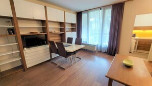 A modern living room with a dining area, built-in shelving, a flat-screen TV, and large windows with curtains.