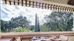 A dining area with a table set for four, a view of a garden with trees and a tall tree, and a striped awning above the window. - Val des Chênes by Interhome (Cavalaire)