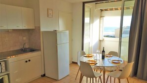 A compact kitchen with a refrigerator, sink, and microwave. There is a dining area with a table set for two, chairs, and a small potted plant. A sofa is visible in the foreground, and the room has a view through a glass door. - Les Balcons de la Méditerranée by Interhome (Narbonne-Plage)