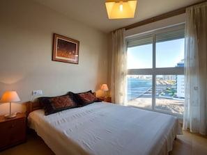 A hotel room with a large bed, two bedside lamps, a framed picture on the wall, and a view of a coastal area through a large window.