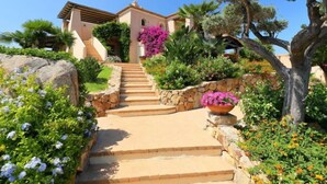 Eine mediterran gestaltete Villa mit einer Steintreppe zum Eingang, umgeben von üppiger Vegetation und blühenden Pflanzen.