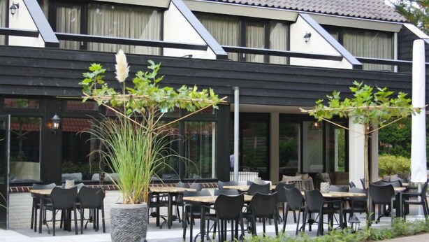An outdoor seating area with tables and chairs, a large planter with plants, and a building with a tiled roof and large windows. - Vacation home 4L by Interhome (Lochem)