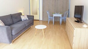 Ein Wohnzimmer mit grauem Sofa, einem kleinen weißen Tisch, einer TV-Konsole und einem Essbereich mit Tisch und Stühlen.