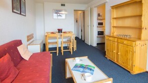 Een knus leefruimte met een rode bank, een houten eettafel met stoelen, een bijzettafeltje met een tijdschrift, en een houten kast met planken.