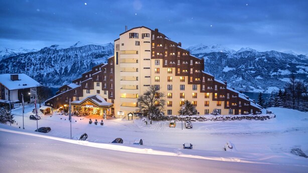Een meerlaags hotel met een skiplaats op de achtergrond, met een opvallende skilift en besneeuwde omgeving.