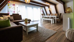 Un loft amplio con techo alto, vigas de madera a la vista, un ventanal con cortinas, un conjunto de sofás, una mesa de comedor y una lámpara colgante.