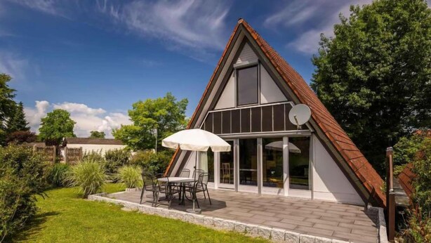 Una casa moderna con techo inclinado, grandes ventanales y un patio con mobiliario exterior.