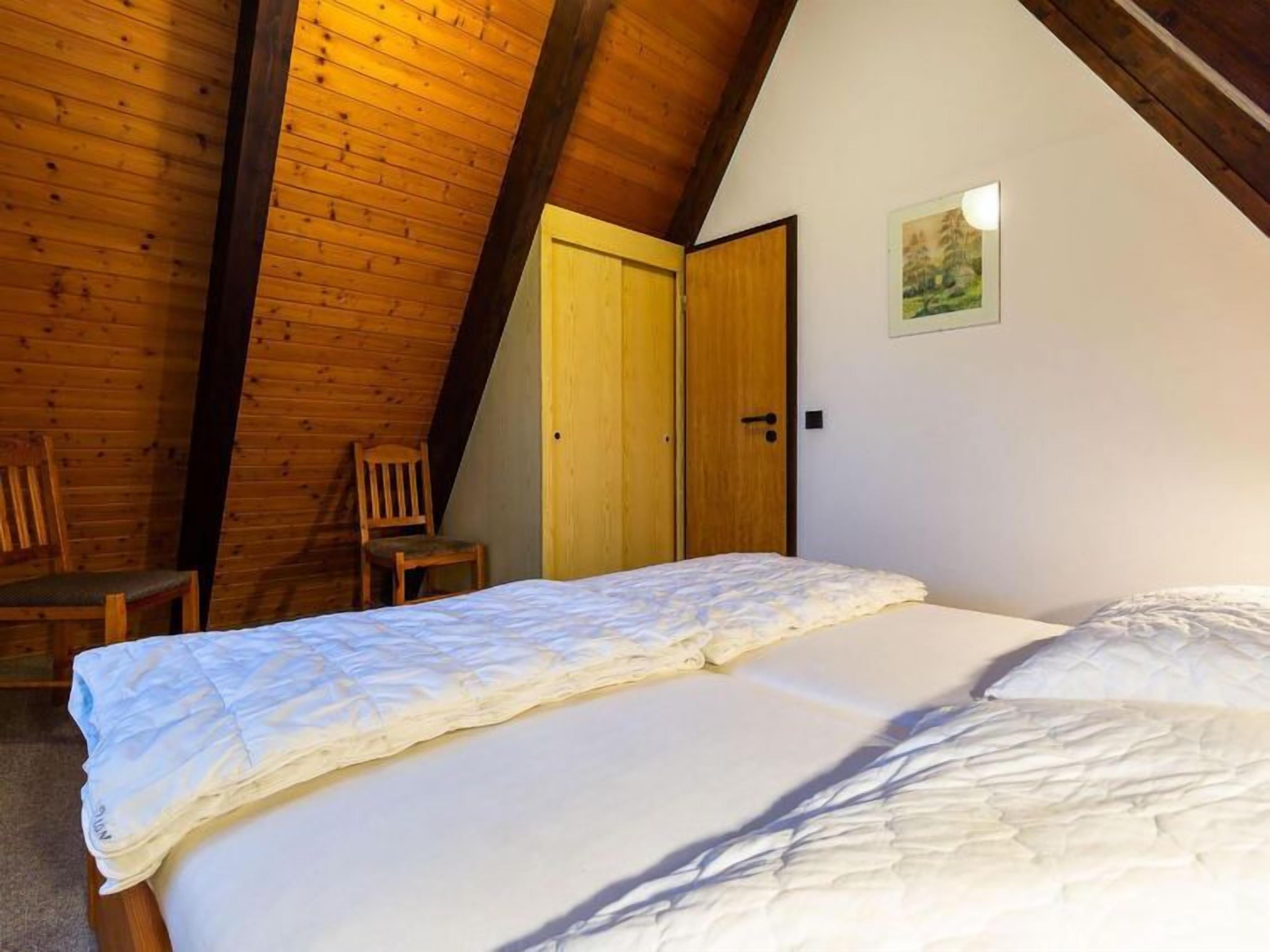 Un dormitorio con techo de madera, una cama con ropa blanca, dos sillas de madera y una puerta.