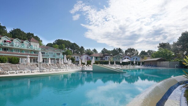 A large outdoor swimming pool with lounge chairs and umbrellas, surrounded by houses with balconies and a clear sky.
