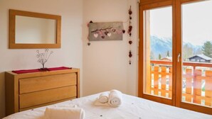 A hotel room with a bed, a wooden dresser, a mirror, a wall decoration, and a view of a wooden deck and mountains through a large window. - Domaine des Reines C10 by Interhome (Nendaz)