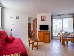 Un salon avec un canapé rouge, un téléviseur posé sur un meuble en bois, un coin repas avec une table et des chaises, et un balcon offrant une vue sur les immebles.