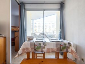 Un coin repas avec une table dressée pour deux personnes, une fenêtre avec des rideaux et une vue sur les bâtiments environnants.
