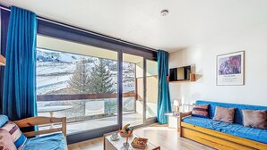 Een moderne woonkamer met een groot raam, een blauwe bank, een houten salontafel en uitzicht op een besneeerd landschap.