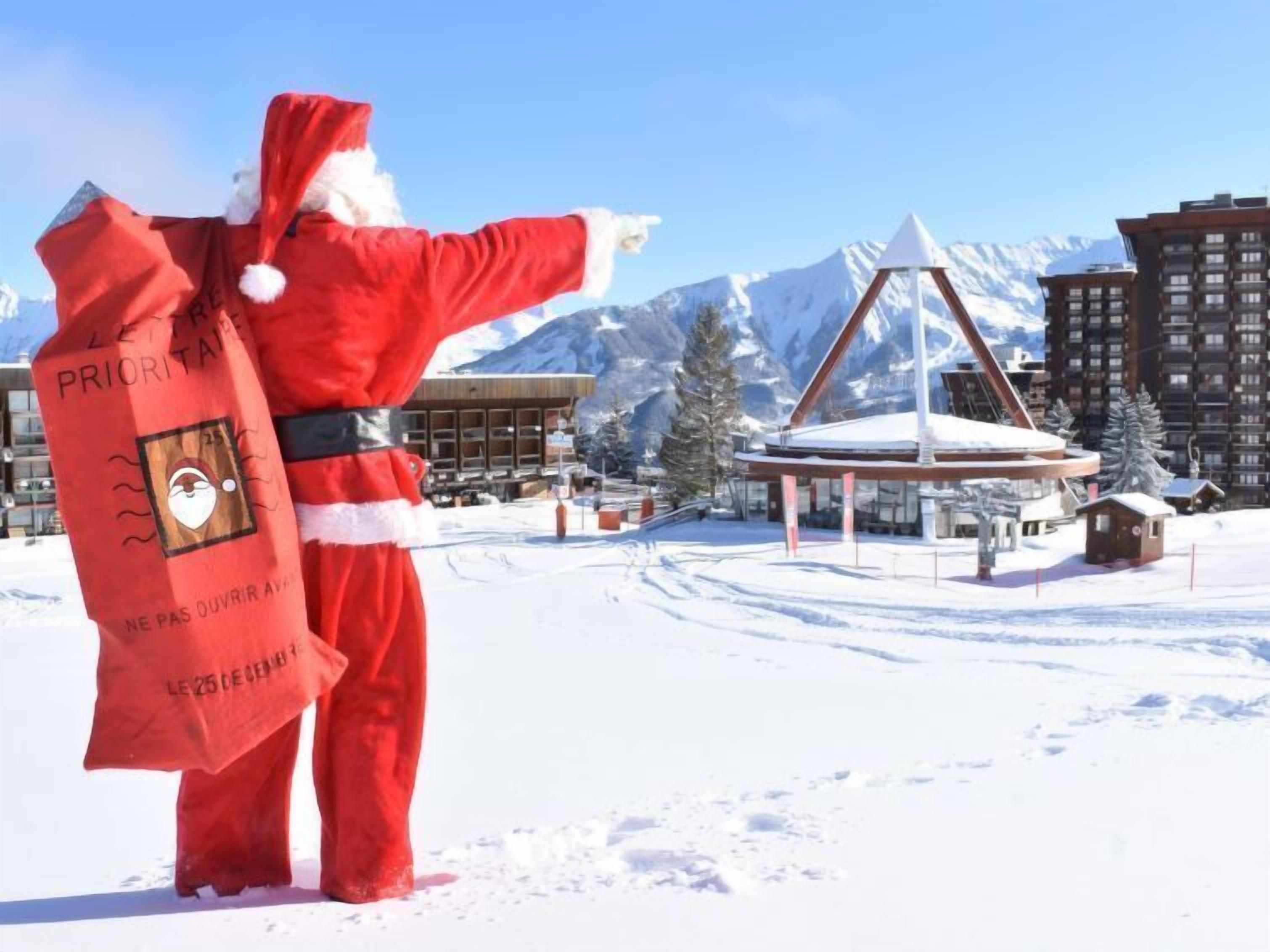 A person in a red suit holding a red bag with text, standing in a snowy area with buildings and mountains in the background.