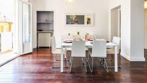Ein Essbereich mit einem weißen Tisch, vier Stühlen und einer Pendelleuchte. Ein Heizkörper und ein Regal mit Dekorationselementen sind ebenfalls vorhanden.