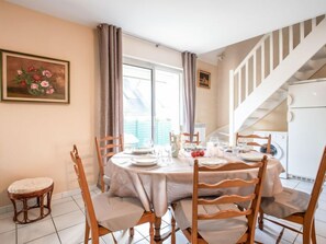 A dining area with a round table set for four, surrounded by wooden chairs. A painting hangs on the wall, and a staircase is visible in the background.
