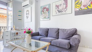 A modern living room with a grey sofa, a glass coffee table, and a white dining table with chairs. There are framed artworks on the wall and a window with a view of the outside. - Le Concordia by Interhome (Nice)