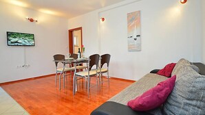 A modern living room with a wooden floor, a black sofa with pink and grey cushions, a dining area with four chairs, a TV mounted on the wall, and two wall-mounted lights. - Apartment Ante by Interhome (Brodarica)