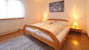 Ein Schlafzimmer mit einem hölzernen Bett, einem großen Fenster mit Vorhängen, einem Blumenbild an der Wand und zwei Nachttischlampen.
