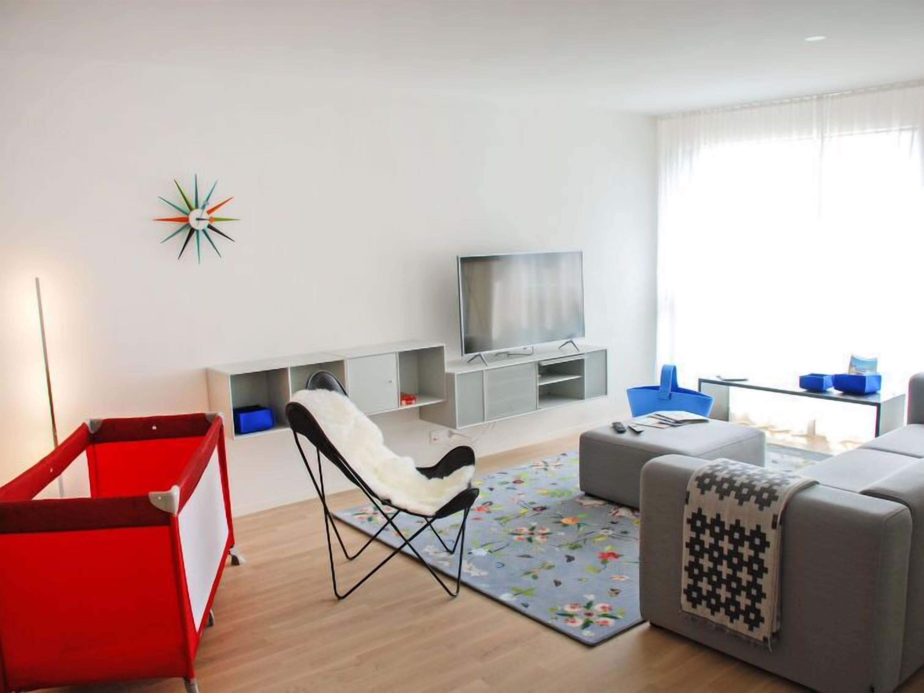 Un soggiorno moderno con una TV a schermo piatto, un recinto gioco rosso, una sedia bianca, un divano grigio e un tappeto floreale.