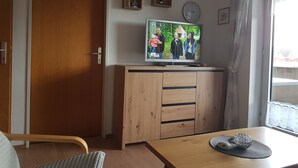 Een kamer met een houten kast, een televisie waarop een gezin te zien is, een klok en een raam met gordijnen.