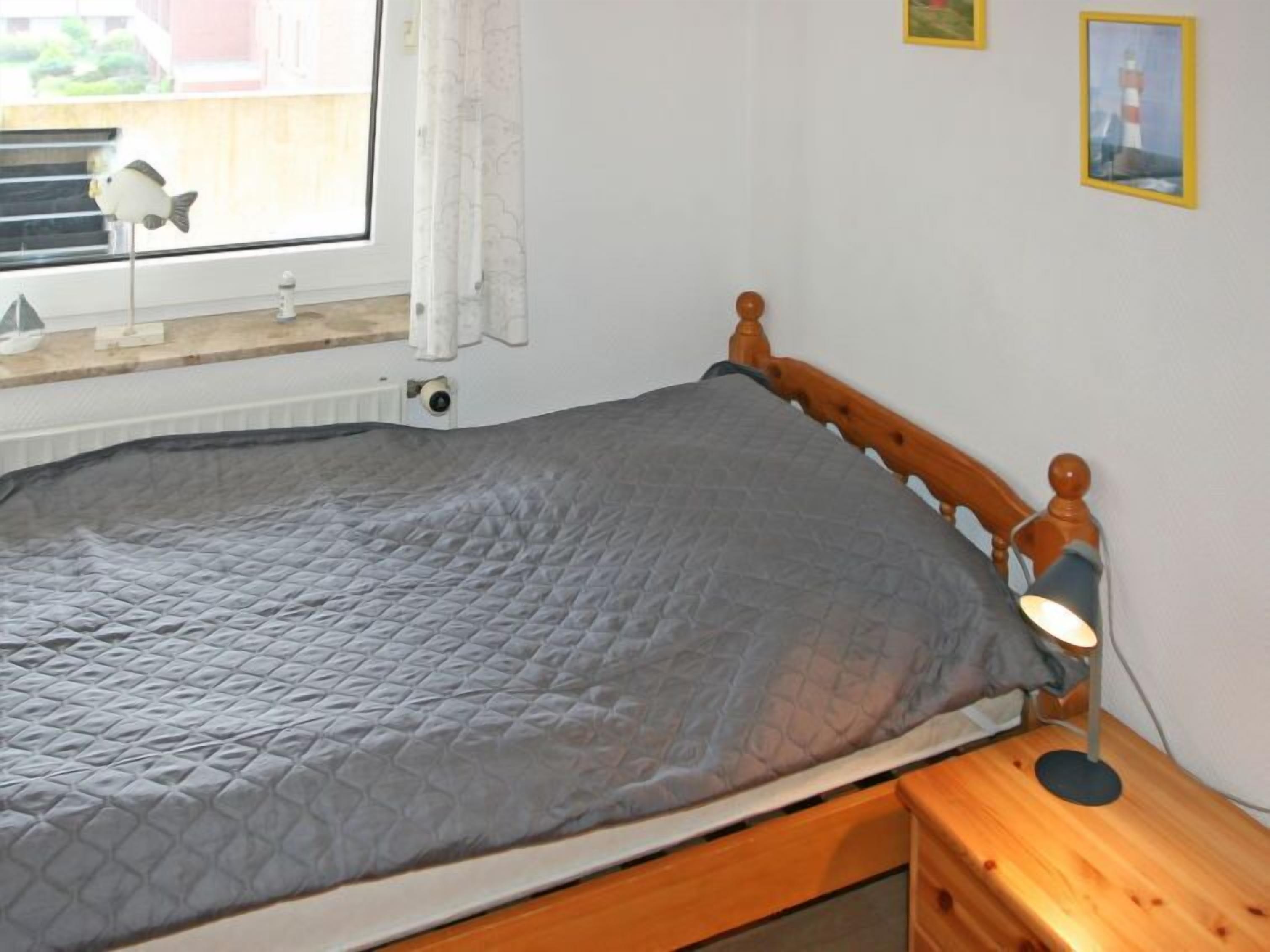 A single bed with a grey quilt, a wooden headboard, a bedside lamp, and a window with a curtain.