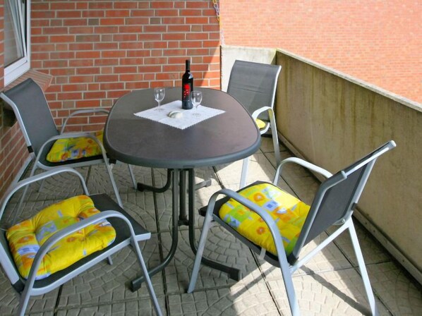 Een klein terras buiten met een ronde tafel en vier stoelen, elk met een geel kussen.