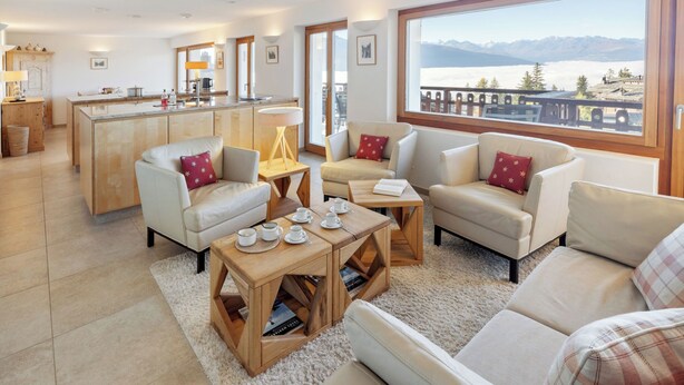A modern living room with a kitchen in the background, a large window with a mountain view, and a cozy seating area with a coffee table.