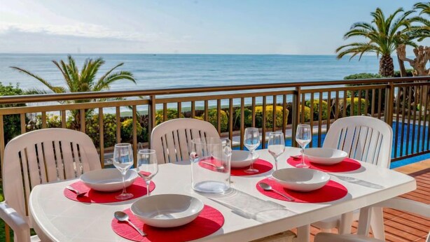 A dining table set for four with white chairs and red placemats, overlooking a pool and the ocean.