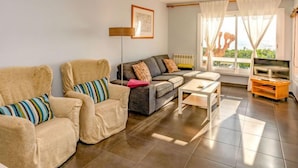 A living room with two beige armchairs, a grey sofa, a white coffee table, and a TV stand. There is a large window with curtains and a view of greenery outside.