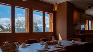 A dining area with a table set for four, large windows with a mountain view, and wooden cabinetry. - Les Chouettes 23 by Interhome (Nendaz)